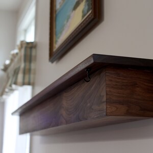 May include: A dark brown wooden shelf with a hidden compartment. The shelf has a hinged lid with a small metal hook on the front. The shelf is mounted on a white wall.