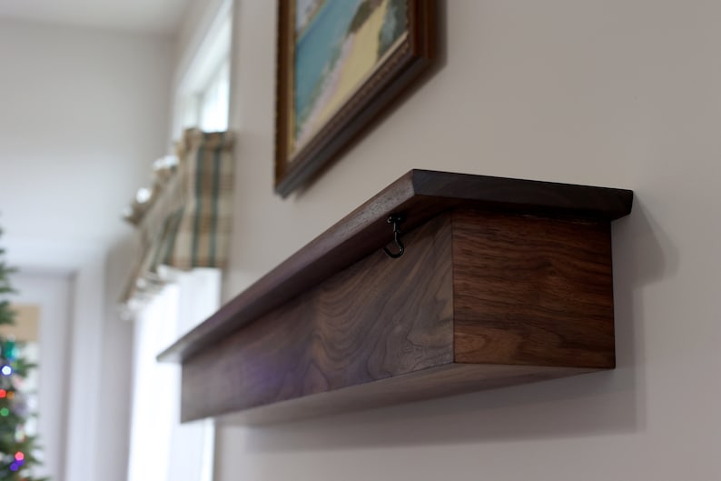 May include: A dark brown wooden shelf with a hidden compartment. The shelf has a hinged lid with a small metal hook on the front. The shelf is mounted on a white wall.