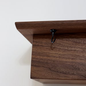 May include: A dark brown wooden shelf with a black metal hook attached to the top. The shelf is made of solid wood and has a natural finish.