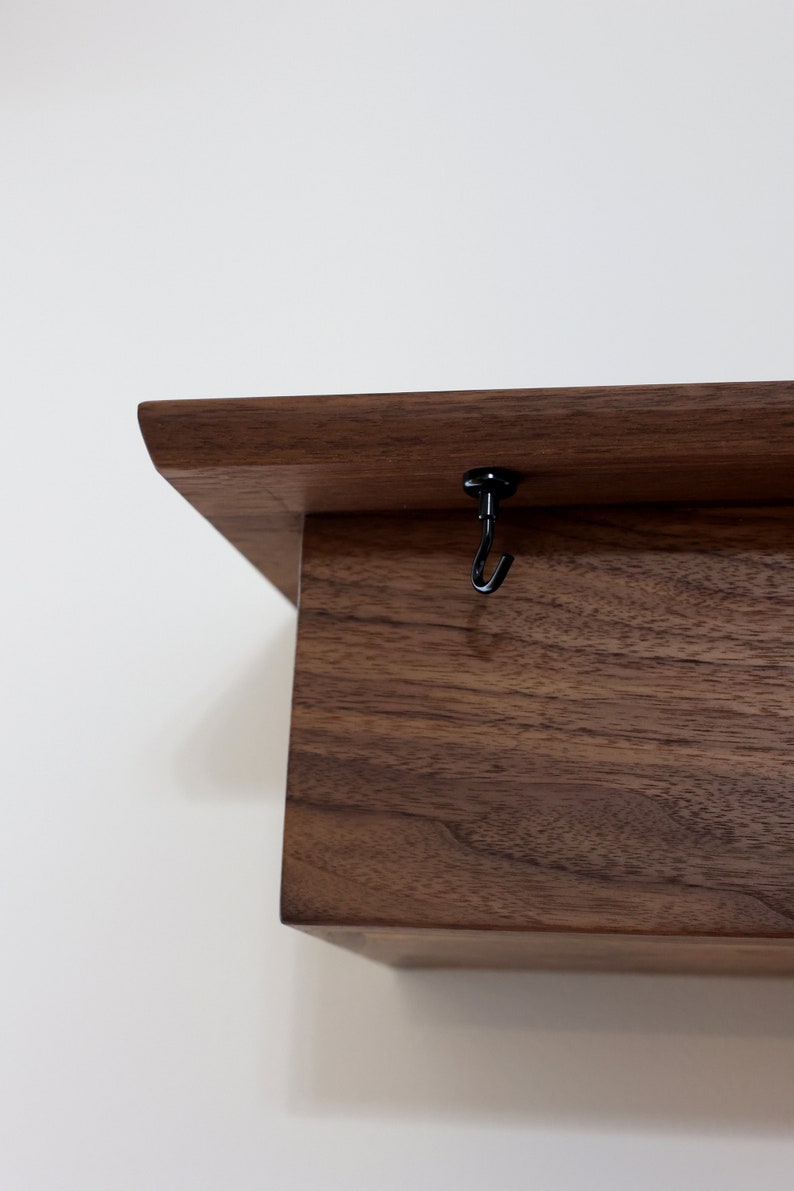 May include: A dark brown wooden shelf with a black metal hook attached to the top. The shelf is made of solid wood and has a natural finish.