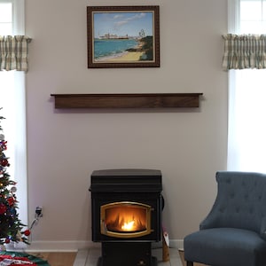 May include: A decorated Christmas tree with colorful lights and ornaments stands next to a window with a plaid valance. A wood mantel shelf is above a black wood-burning stove with a roaring fire. A blue armchair sits in the corner of the room.