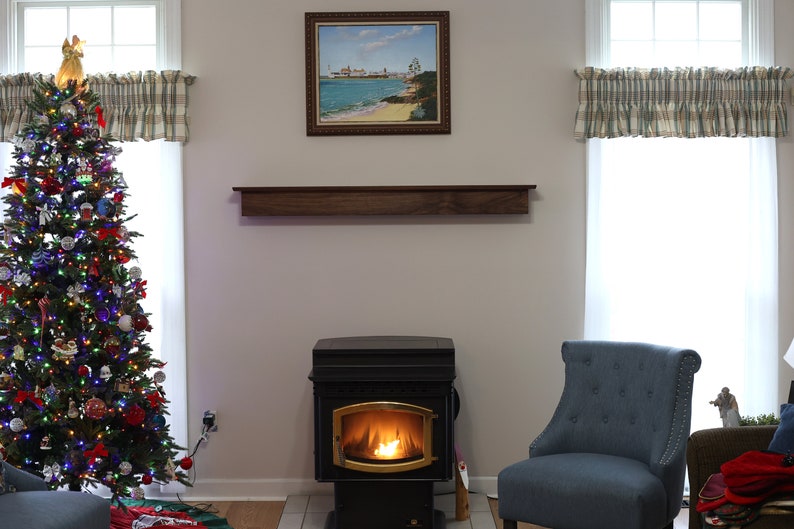 May include: A decorated Christmas tree with colorful lights and ornaments stands next to a window with a plaid valance. A wood mantel shelf is above a black wood-burning stove with a roaring fire. A blue armchair sits in the corner of the room.