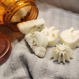 May include: Five white, handmade soap bars in various shapes, including a flower, a star, and a horseshoe. The soap bars have a light brown, herbal scent and are arranged on a white towel. The soap bars are packaged in a clear glass jar with a brown lid. The lid has a sticker that says "handmade with love".