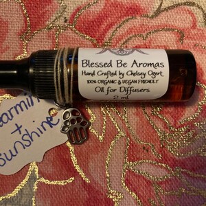 May include: A small brown glass bottle with a dropper top and a white label that reads "Blessed Be Aromas Hand Crafted by Chelsey Ogert 100% Organic & Vegan Friendly Oil for Diffusers 2 ml". The bottle is sitting on a pink and gold fabric with a white tag that reads "Spearmint + Sunshine".