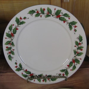 May include: White ceramic plate with a green and red holly berry design around the rim. The plate has a dark blue border. The plate is displayed on a decorative metal stand. A festive holiday theme.