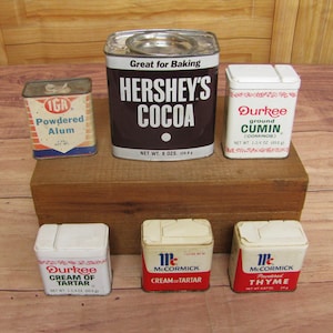 May include: A collection of vintage spice tins, including a tin of Hershey's Cocoa, a tin of Durkee Ground Cumin, a tin of Durkee Cream of Tartar, and two tins of McCormick Cream of Tartar and McCormick Thyme. The tins are arranged on a wooden surface.
