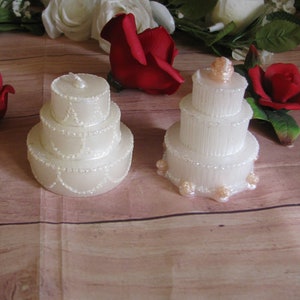 May include: Two white wedding cake candles with pearl accents. The larger candle has three tiers and the smaller candle has two tiers. Both candles have a small rose on top.