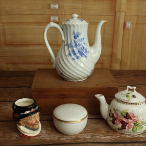 May include: A collection of antique porcelain teaware. Includes a white and blue swirl-patterned coffee pot, a small teapot with floral designs, a small lidded container, and a novelty mug with a man's face.