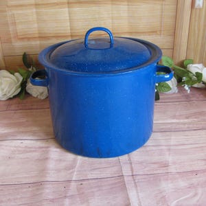 May include: A deep, cylindrical stockpot with a vibrant blue enamel finish and a speckled white pattern. The pot features two side handles and a matching lid with a central loop handle, providing a sturdy design for cooking large quantities.
