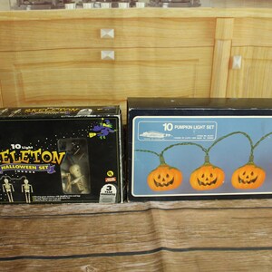 May include: Two rectangular cardboard boxes containing Halloween light sets. The left box is black with the text 10 Light SKELETON HALLOWEEN SET and features skeleton graphics. The right box is blue with the text 10 PUMPKIN LIGHT SET.