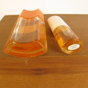 May include: Two vintage perfume bottles. One is a tall, angular bottle with an orange cap. The other is a smaller, cylindrical bottle with a white cap. Both bottles contain a light amber liquid. The bottles are on a wooden surface.