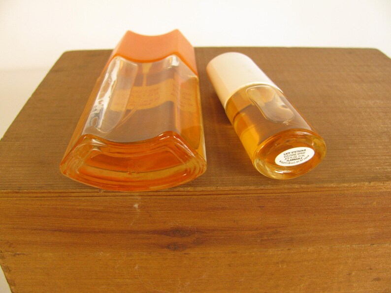 May include: Two vintage perfume bottles. One is a tall, angular bottle with an orange cap. The other is a smaller, cylindrical bottle with a white cap. Both bottles contain a light amber liquid. The bottles are on a wooden surface.