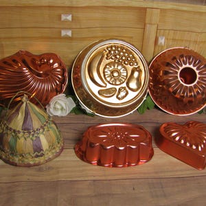 May include: A collection of five metallic copper-toned baking molds in various shapes including a shell, a fruit-themed round mold, a fluted ring, an oval floral mold, and a heart, alongside a woven conical food cover with purple and green accents.