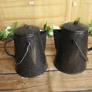 May include: Two black enamel coffee pots with white speckled patterns. Each pot features a tapered cylindrical body, a matching lid with a small knob, a side handle, and a metal wire bail handle attached to the sides for carrying or hanging.
