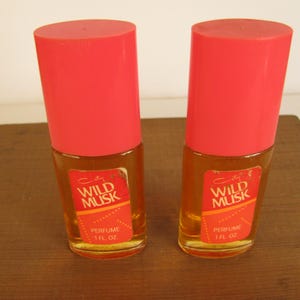 May include: Two vintage perfume bottles with bright red caps. The bottles have a rectangular label with the words "Wild Musk" and "Perfume 1 fl oz". The perfume inside is amber-colored.