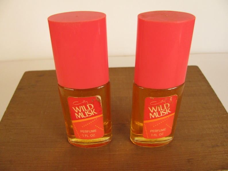 May include: Two vintage perfume bottles with bright red caps. The bottles have a rectangular label with the words "Wild Musk" and "Perfume 1 fl oz". The perfume inside is amber-colored.