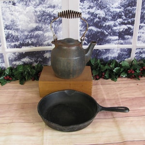 May include: A vintage cast iron teapot with a spring handle and a matching cast iron skillet. Both pieces are dark gray with signs of rust.