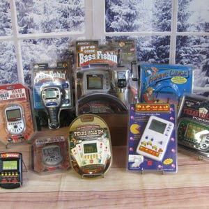 May include: A collection of handheld electronic games, including fishing, poker, and Pac-Man. The games are packaged in clear plastic boxes with colorful graphics and text.