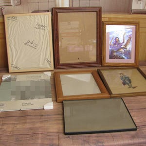 May include: A collection of seven rectangular picture frames in various materials and finishes, including light wood, dark wood, and black metal. Some frames contain glass, while others hold prints of a person with an eagle or a seated figure.