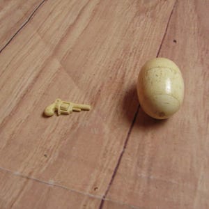 May include: A small, white plastic toy gun and a white egg-shaped object on a wooden surface.
