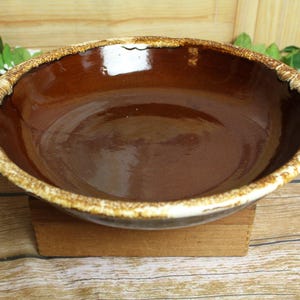 May include: A brown ceramic bowl with a glossy finish. The rim has a textured, off-white and brown edge. The bowl is set on a wooden block, with a wooden background and floral accents.