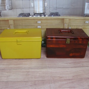 May include: Two storage boxes, one yellow and one brown. The yellow box is made of plastic and has a handle on the top. The brown box is made of clear plastic and has a metal latch.