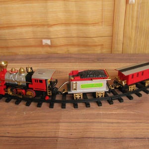May include: A toy train set with a red, silver, and black locomotive, a coal car, and a red caboose on a black track. The coal car has the text "Christmas Express". The train is on a wooden surface.