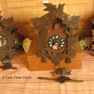 May include: Three brown cuckoo clocks with dark brown leaf and bird accents. The clocks have black clock faces with white Roman numerals. The clocks are on a wooden surface. The text "2-Two Tone Clock" is at the bottom.