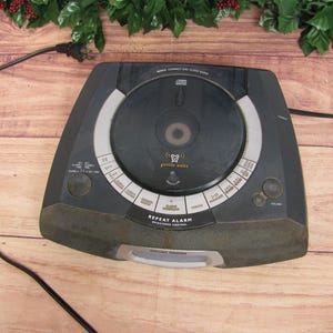 May include: Black CD clock radio with a silver face and a large round CD player. The radio has a digital display and a variety of buttons for setting the time, alarm, and other functions. The text on the radio says "REPEAT ALARM" and "GENTLE WAKE".