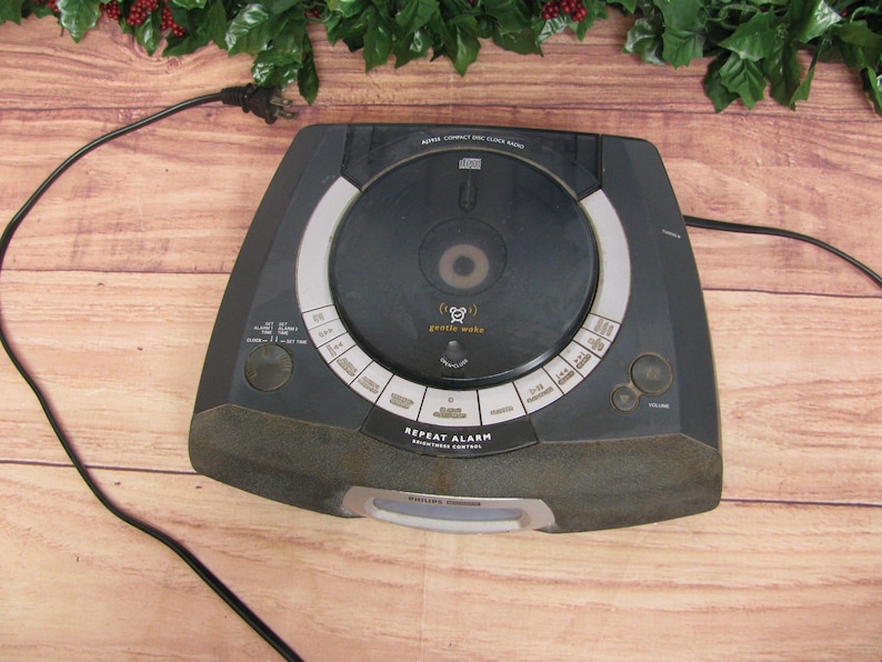 May include: Black CD clock radio with a silver face and a large round CD player. The radio has a digital display and a variety of buttons for setting the time, alarm, and other functions. The text on the radio says "REPEAT ALARM" and "GENTLE WAKE".
