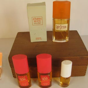 May include: An assortment of vintage perfume bottles and a box are displayed on a wooden surface. The bottles have orange and red caps, with labels that read "Jovan White Musk", "Lady Stetson", and "Wild Musk". The box is labeled "Jovan White Musk".