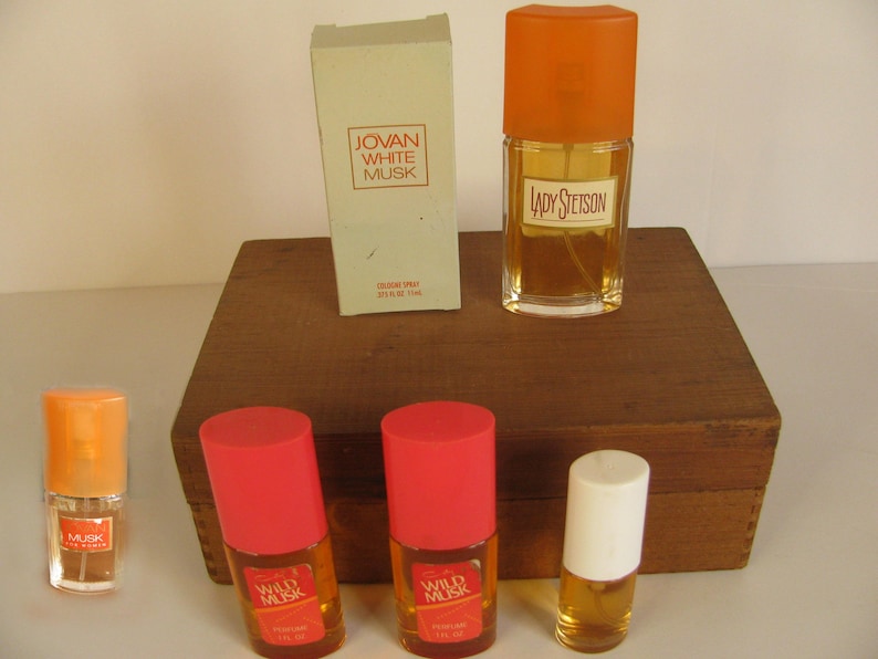 May include: An assortment of vintage perfume bottles and a box are displayed on a wooden surface. The bottles have orange and red caps, with labels that read "Jovan White Musk", "Lady Stetson", and "Wild Musk". The box is labeled "Jovan White Musk".