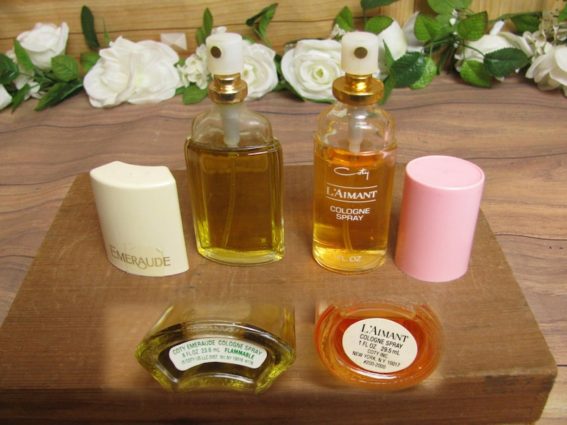 May include: An assortment of vintage perfume bottles and cologne sprays. The bottles are filled with amber-colored liquid. One bottle is labeled "L'Aimant Cologne Spray" and another "Emeraude". A pink cap and a white cap are also present.