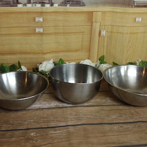 May include: Three polished stainless steel mixing bowls of varying sizes arranged in a horizontal row. Each bowl features a smooth, rounded interior and a simple, clean design with a reflective metallic finish, suitable for kitchen preparation tasks.