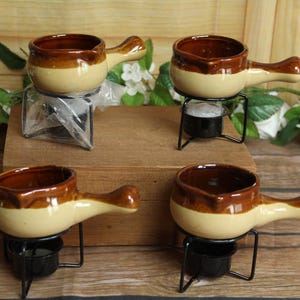 May include: A set of four ceramic fondue pots with a two-tone cream and brown glaze, each featuring a single handle and a pouring spout. Each pot rests on a black metal wire stand designed to hold a small tea light candle underneath the base.