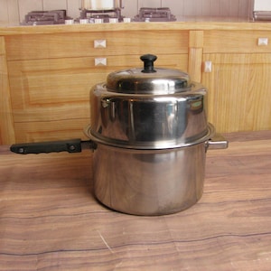 May include: Stainless steel steamer pot with a black handle and lid. The pot has a round base and a steamer insert. The lid has a black knob. The pot is sitting on a wooden surface.