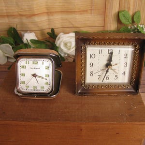May include: Two vintage alarm clocks, one silver with a square face and the other gold with a round face. Both clocks have black hands and Roman numeral markings.