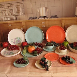 May include: A set of vintage melamine dinnerware in various colors, including blue, orange, and white. The plates feature a speckled pattern and are arranged on a wooden table with grapes and apples.