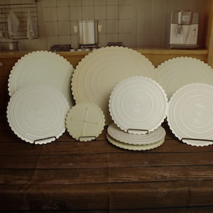 May include: A collection of round, off-white cake boards with scalloped edges. The set includes various sizes, featuring concentric circular embossed patterns on the surface. Some boards are stacked, while others stand upright, displaying a consistent light cream color and a textured, decorative border design.