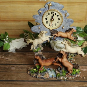 May include: A decorative clock featuring a light blue tree-shaped frame with a clock face. The clock has a detailed base with several brown and white horse figurines. The clock is set against a wooden background.