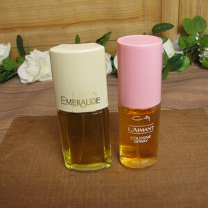 May include: Two vintage perfume bottles on a wooden surface. One bottle has a cream-colored cap with "Emeraude" printed on it. The other has a pink cap and the text "L'Aimant Cologne Spray". Both bottles contain a golden liquid.