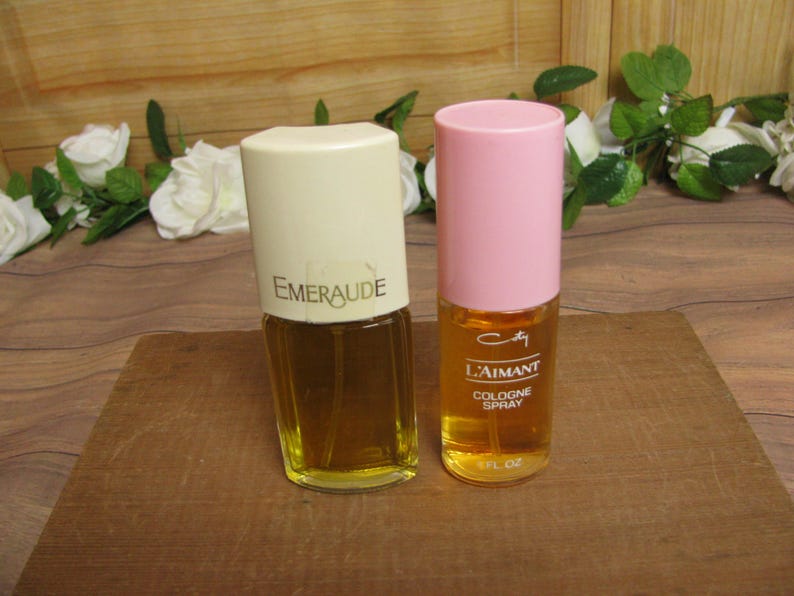 May include: Two vintage perfume bottles on a wooden surface. One bottle has a cream-colored cap with "Emeraude" printed on it. The other has a pink cap and the text "L'Aimant Cologne Spray". Both bottles contain a golden liquid.