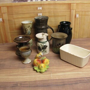 May include: A collection of ceramic pottery in various shapes and sizes, including vases, bowls, and a rectangular planter. The pottery features a range of colors, from dark brown to cream, with some pieces displaying decorative patterns. A small ceramic fruit sculpture is also present.