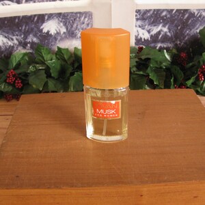 May include: A small perfume bottle with an orange cap and a label that reads "Swan Musk for Women." The bottle is clear, and the liquid inside is a light amber color. The bottle is on a wooden surface.