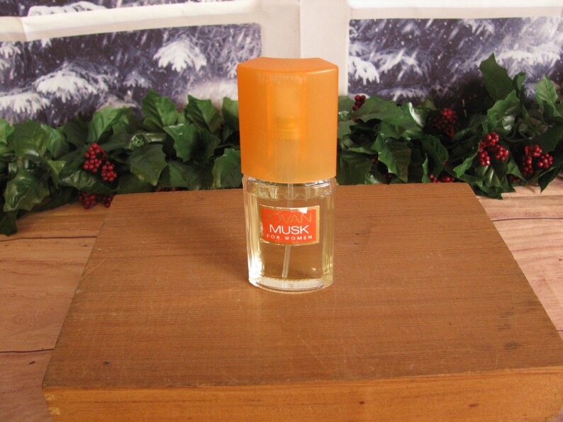 May include: A small perfume bottle with an orange cap and a label that reads "Swan Musk for Women." The bottle is clear, and the liquid inside is a light amber color. The bottle is on a wooden surface.