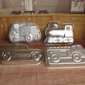 May include: Four silver-colored metal cake pans in the shapes of a tractor, train, SUV, and fire truck. The pans are arranged on a wooden surface, showcasing their detailed designs and intended use for baking themed cakes.