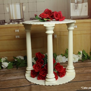 May include: A white tiered cake stand featuring four fluted columns supporting a circular top platform. The stand is decorated with vibrant red roses placed on the top surface and between the columns on the base. The structure is freestanding.