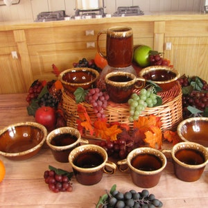 May include: A set of brown and beige glazed ceramic mugs and bowls. The mugs and bowls have a rustic, vintage look. The set is arranged on a wicker basket with grapes and fall leaves.