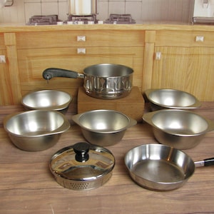 May include: A collection of miniature stainless steel cookware, including a saucepan, frying pan, bowls, and a lidded steamer. The set is arranged on a wooden surface, suggesting a kitchen or play set theme.