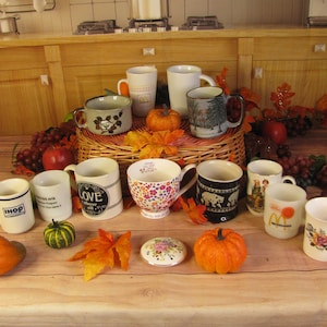 May include: A collection of 11 ceramic mugs with various designs and colors. The mugs are arranged on a wooden table with fall decorations, including pumpkins, grapes, and leaves.